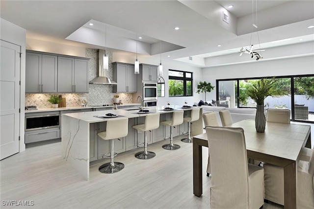 kitchen featuring gray cabinets, a raised ceiling, hanging light fixtures, a kitchen island with sink, and wall chimney exhaust hood