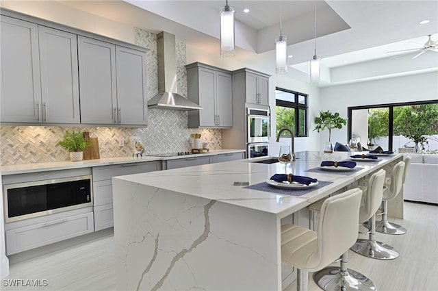 kitchen featuring gray cabinets, a kitchen island with sink, built in microwave, wall chimney exhaust hood, and light stone counters