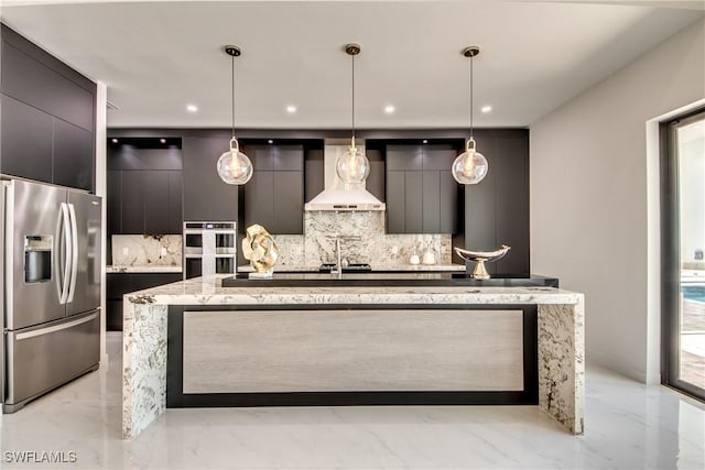 kitchen with decorative light fixtures, an island with sink, backsplash, stainless steel appliances, and wall chimney range hood