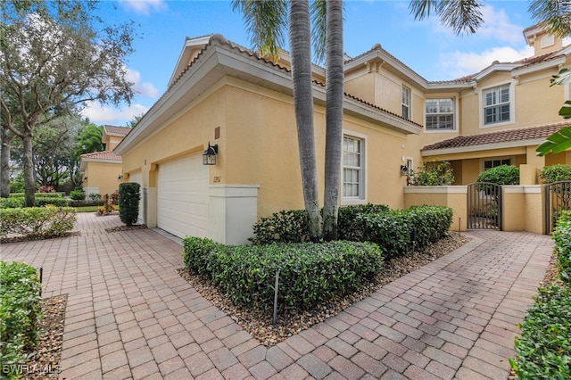 view of side of home featuring a tile roof, stucco siding, decorative driveway, an attached garage, and a gate