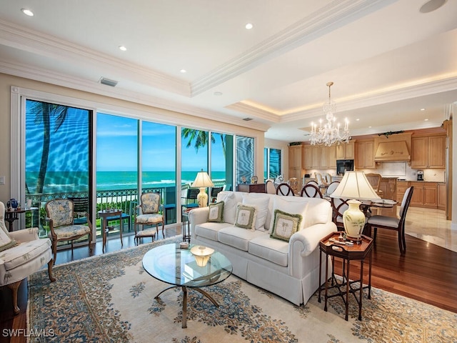 living room featuring a notable chandelier, crown molding, a tray ceiling, a water view, and light wood-type flooring