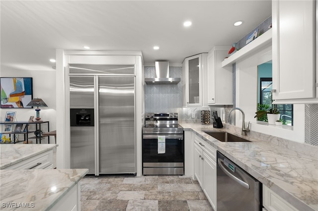 kitchen featuring white cabinets, appliances with stainless steel finishes, light stone countertops, wall chimney range hood, and a sink