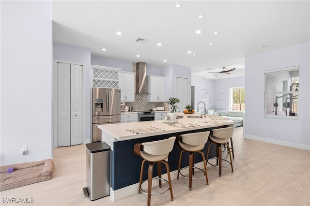 kitchen with wall chimney exhaust hood, stainless steel appliances, a large island with sink, a kitchen bar, and white cabinets