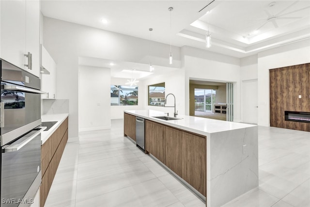 kitchen with sink, a raised ceiling, an island with sink, decorative light fixtures, and white cabinets