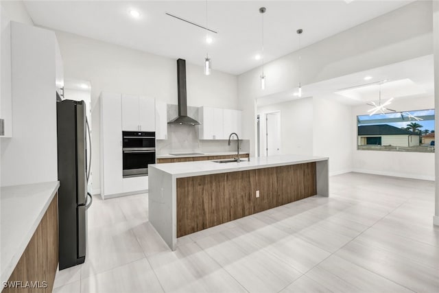 kitchen with a kitchen island with sink, wall chimney range hood, black appliances, decorative light fixtures, and white cabinets