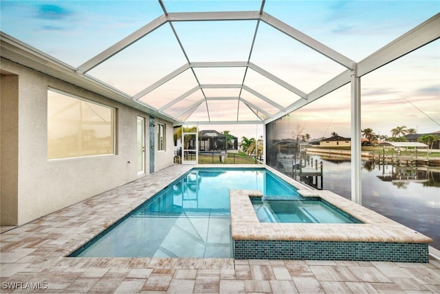 pool at dusk with glass enclosure, a patio area, a water view, and an in ground hot tub