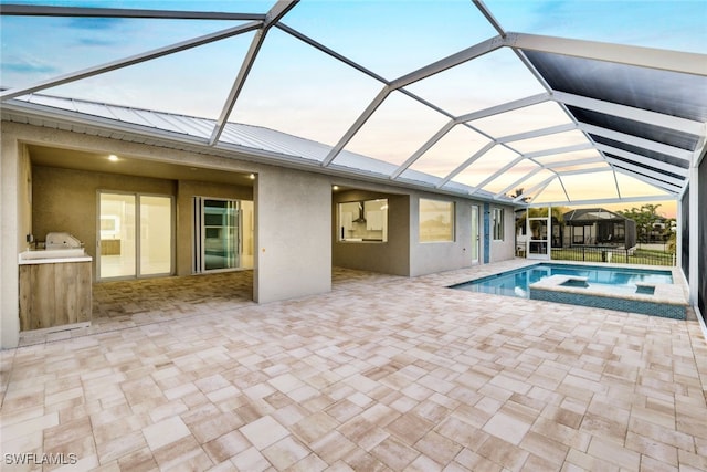 pool at dusk featuring glass enclosure, a patio area, and an in ground hot tub