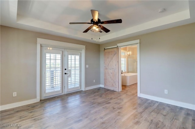 unfurnished room featuring french doors, a raised ceiling, light hardwood / wood-style flooring, ceiling fan, and a barn door