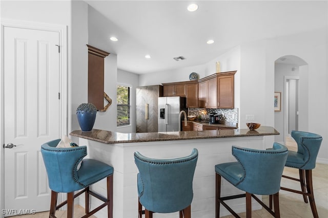 kitchen with kitchen peninsula, stainless steel refrigerator with ice dispenser, backsplash, light tile patterned flooring, and dark stone counters