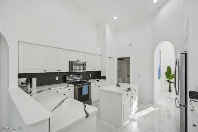 kitchen featuring a towering ceiling, backsplash, stainless steel appliances, white cabinets, and a kitchen island