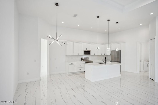 kitchen featuring a towering ceiling, an island with sink, decorative light fixtures, white cabinets, and appliances with stainless steel finishes