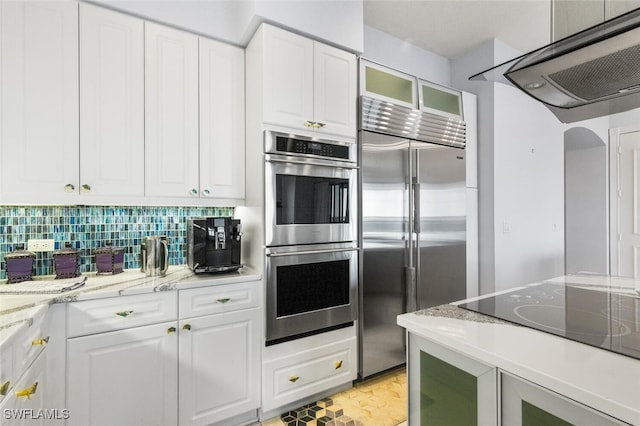 kitchen with appliances with stainless steel finishes, exhaust hood, white cabinetry, tasteful backsplash, and light stone counters