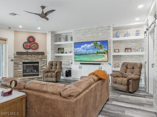 living room featuring ceiling fan, a barn door, light wood-type flooring, a stone fireplace, and built in shelves