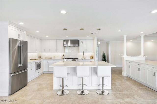 kitchen featuring decorative light fixtures, an island with sink, white cabinets, stainless steel appliances, and wall chimney exhaust hood