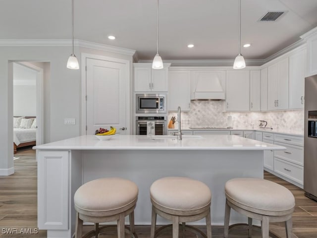 kitchen with hanging light fixtures, stainless steel appliances, a center island with sink, custom range hood, and white cabinets