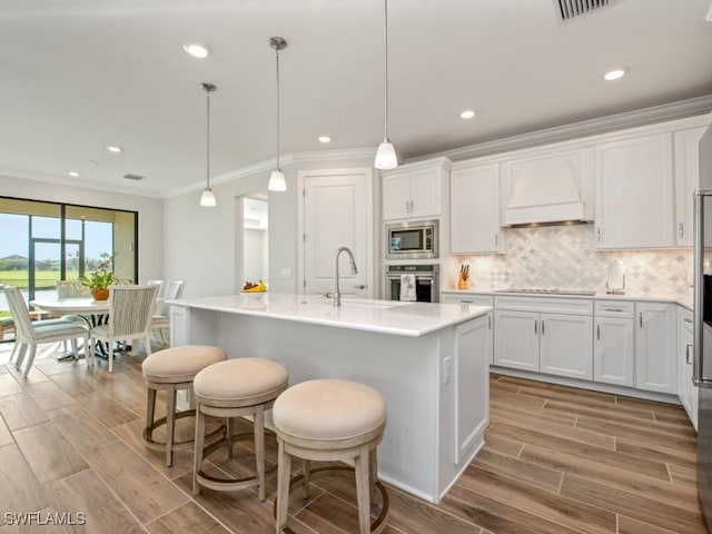 kitchen with white cabinetry, decorative light fixtures, a kitchen island with sink, and premium range hood