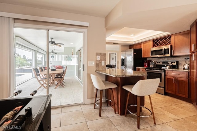 kitchen with a kitchen bar, stainless steel appliances, kitchen peninsula, a raised ceiling, and light stone counters