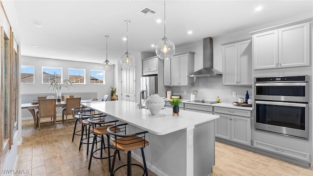 kitchen featuring stainless steel appliances, hanging light fixtures, wall chimney range hood, a breakfast bar area, and a kitchen island with sink