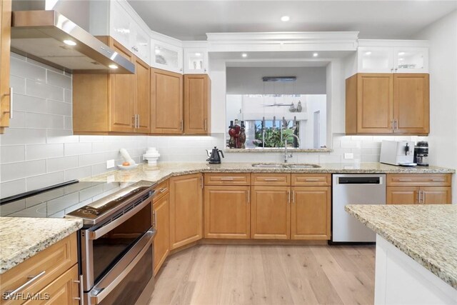 kitchen with wall chimney range hood, sink, stainless steel appliances, light stone counters, and light hardwood / wood-style floors