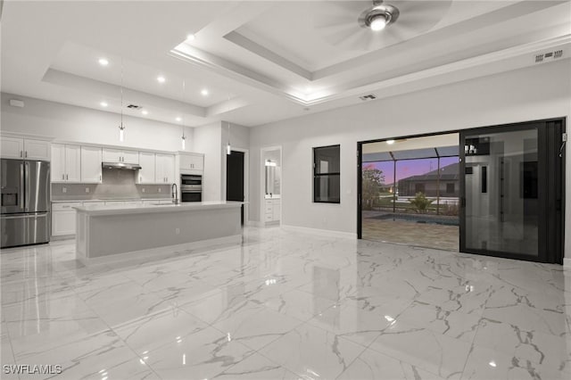 kitchen featuring stainless steel appliances, an island with sink, white cabinets, backsplash, and a tray ceiling