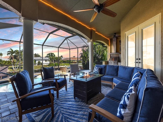 patio terrace at dusk featuring a lanai, an outdoor living space with a fire pit, an outdoor kitchen, and french doors