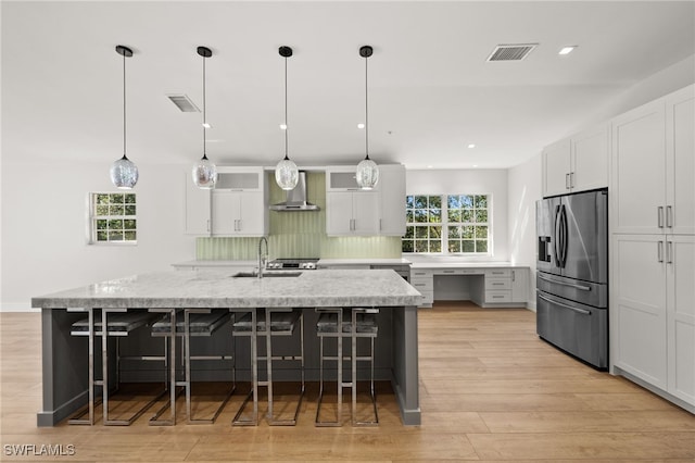 kitchen featuring stainless steel refrigerator with ice dispenser, wall chimney exhaust hood, sink, decorative light fixtures, and white cabinetry