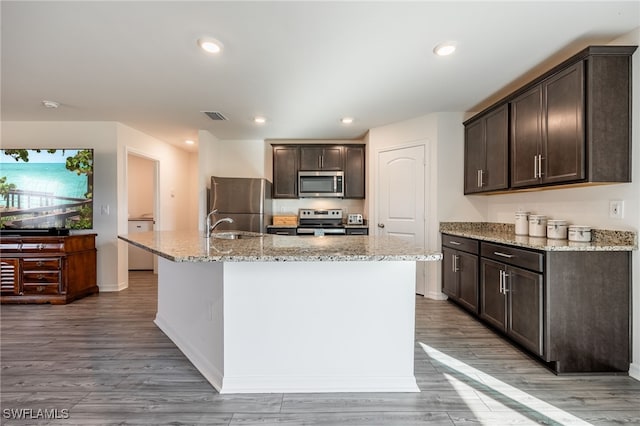 kitchen featuring light stone countertops, light hardwood / wood-style floors, a kitchen island with sink, dark brown cabinets, and appliances with stainless steel finishes