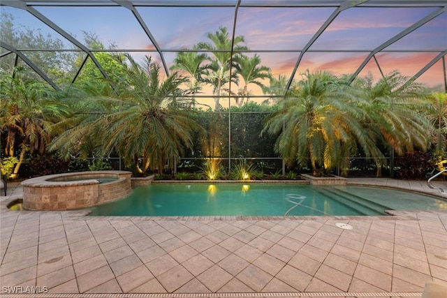 pool at dusk featuring glass enclosure, an in ground hot tub, and a patio