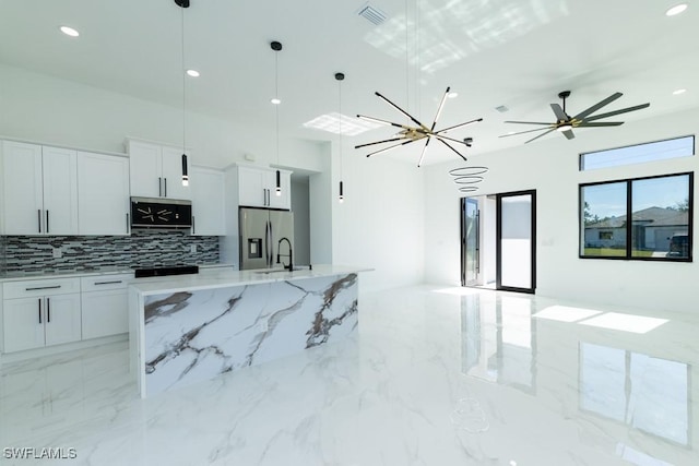 kitchen featuring white cabinetry, hanging light fixtures, stainless steel refrigerator with ice dispenser, light stone counters, and a large island with sink