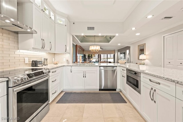 kitchen with stainless steel appliances, decorative backsplash, a raised ceiling, wall chimney exhaust hood, and white cabinets