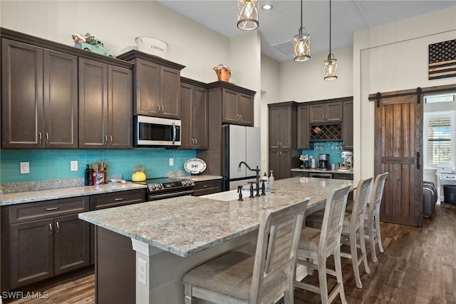 kitchen featuring appliances with stainless steel finishes, dark wood-type flooring, a barn door, decorative light fixtures, and an island with sink