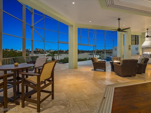 patio terrace at dusk featuring ceiling fan, outdoor lounge area, a water view, a grill, and exterior kitchen