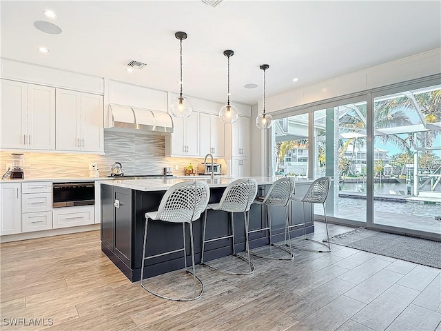 kitchen with white cabinetry, an island with sink, pendant lighting, light hardwood / wood-style floors, and decorative backsplash