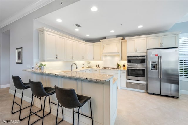 kitchen featuring stainless steel appliances, light stone counters, premium range hood, kitchen peninsula, and a kitchen bar