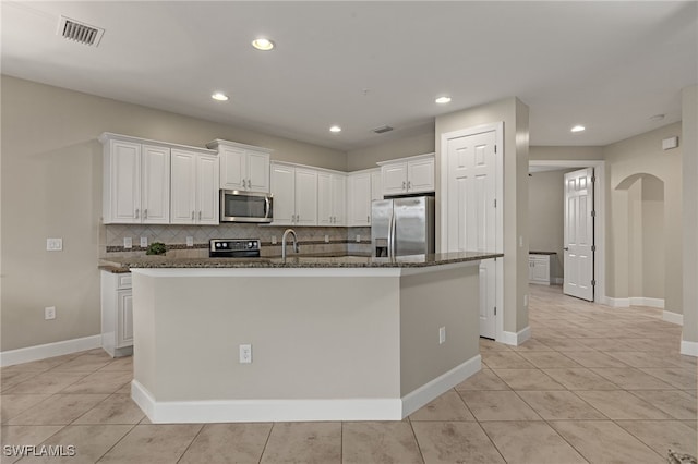 kitchen featuring white cabinetry, an island with sink, appliances with stainless steel finishes, backsplash, and dark stone counters