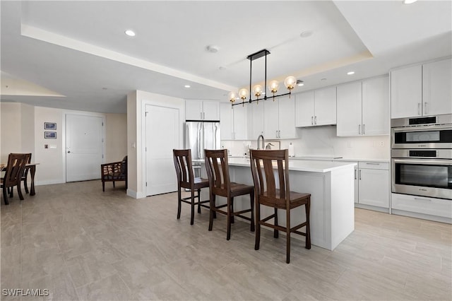 kitchen featuring stainless steel appliances, white cabinetry, a raised ceiling, hanging light fixtures, and a kitchen island with sink