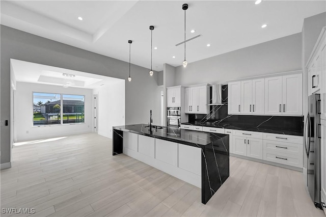 kitchen with white cabinetry, wall chimney range hood, decorative light fixtures, a center island with sink, and appliances with stainless steel finishes