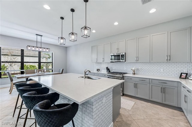 kitchen with gray cabinetry, sink, stainless steel appliances, pendant lighting, and a center island with sink