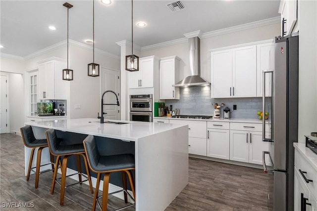 kitchen featuring wall chimney range hood, hanging light fixtures, sink, an island with sink, and appliances with stainless steel finishes