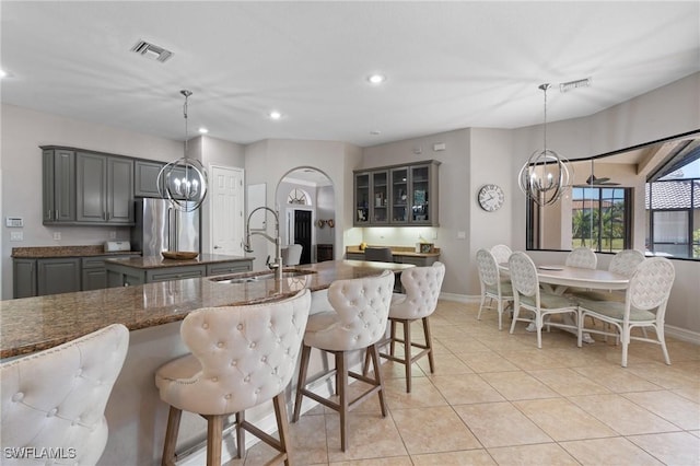 kitchen with gray cabinets, an island with sink, dark stone counters, pendant lighting, and stainless steel fridge