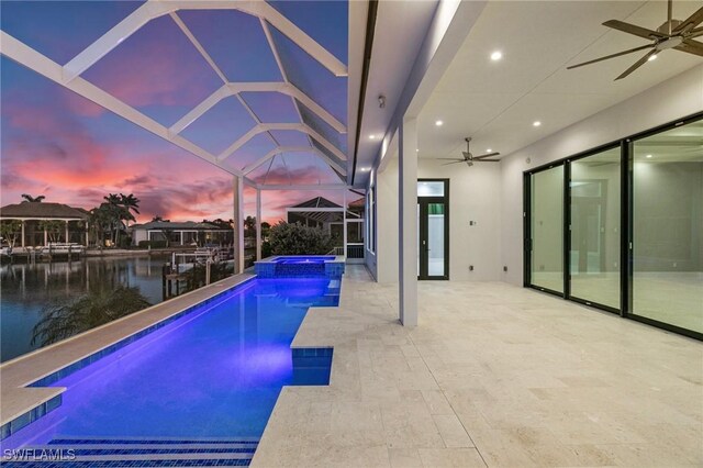 pool at dusk featuring a lanai, ceiling fan, a patio area, a water view, and an in ground hot tub