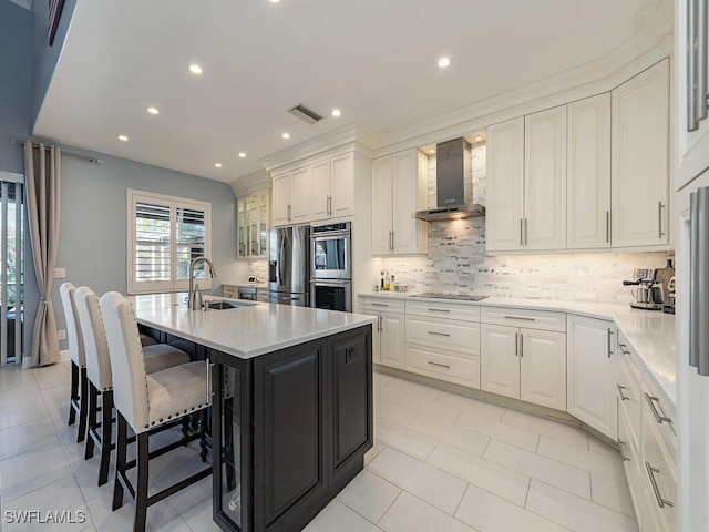 kitchen with stainless steel appliances, sink, wall chimney range hood, white cabinets, and an island with sink