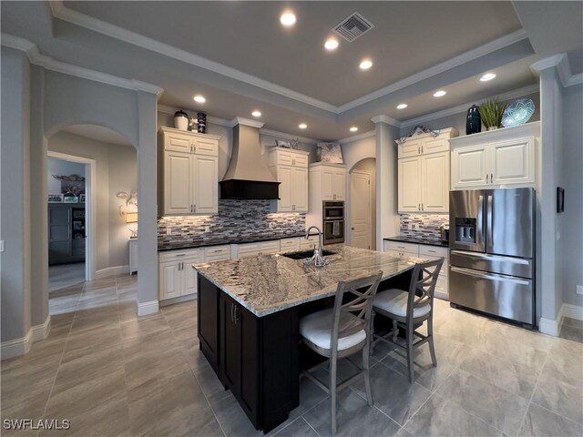 kitchen featuring appliances with stainless steel finishes, custom exhaust hood, an island with sink, sink, and light stone counters