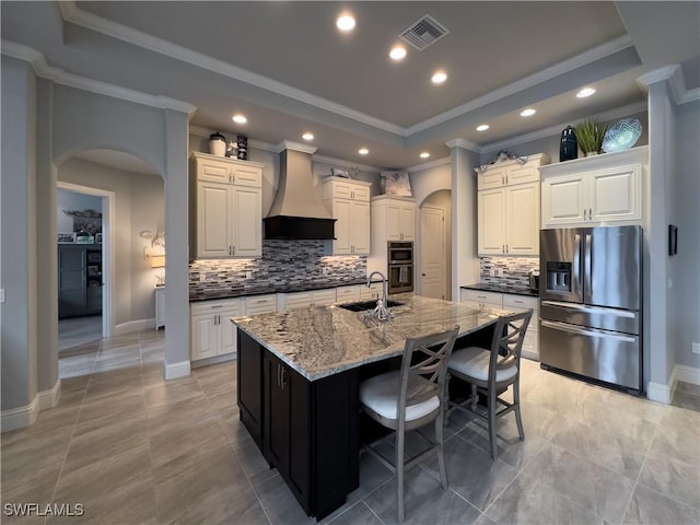 kitchen with a tray ceiling, stainless steel appliances, premium range hood, and a center island with sink