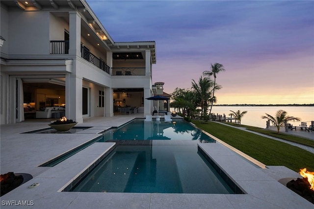 pool at dusk with an outdoor fire pit, a patio area, a lawn, and a water view