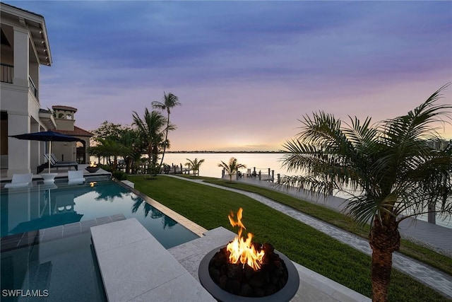 pool at dusk with an outdoor fire pit, a lawn, and a water view