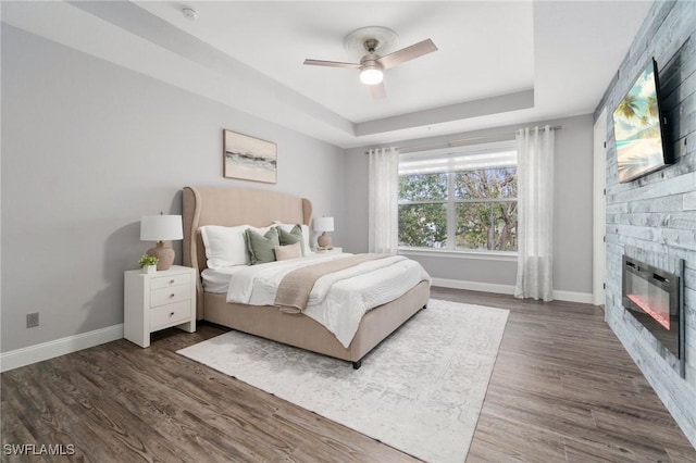 bedroom featuring dark wood-type flooring, ceiling fan, a raised ceiling, and a stone fireplace