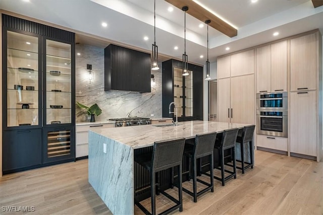 kitchen with a kitchen island with sink, pendant lighting, light stone counters, beam ceiling, and exhaust hood