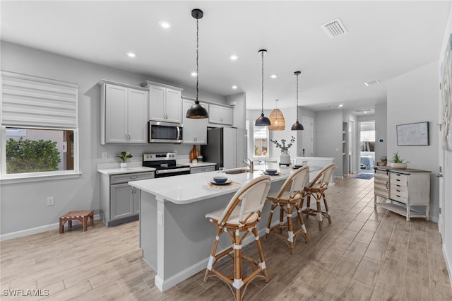 kitchen featuring decorative light fixtures, gray cabinets, a kitchen island with sink, a breakfast bar, and appliances with stainless steel finishes