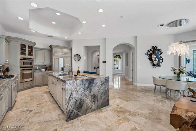 kitchen with gray cabinets, an island with sink, light stone countertops, a tray ceiling, and sink